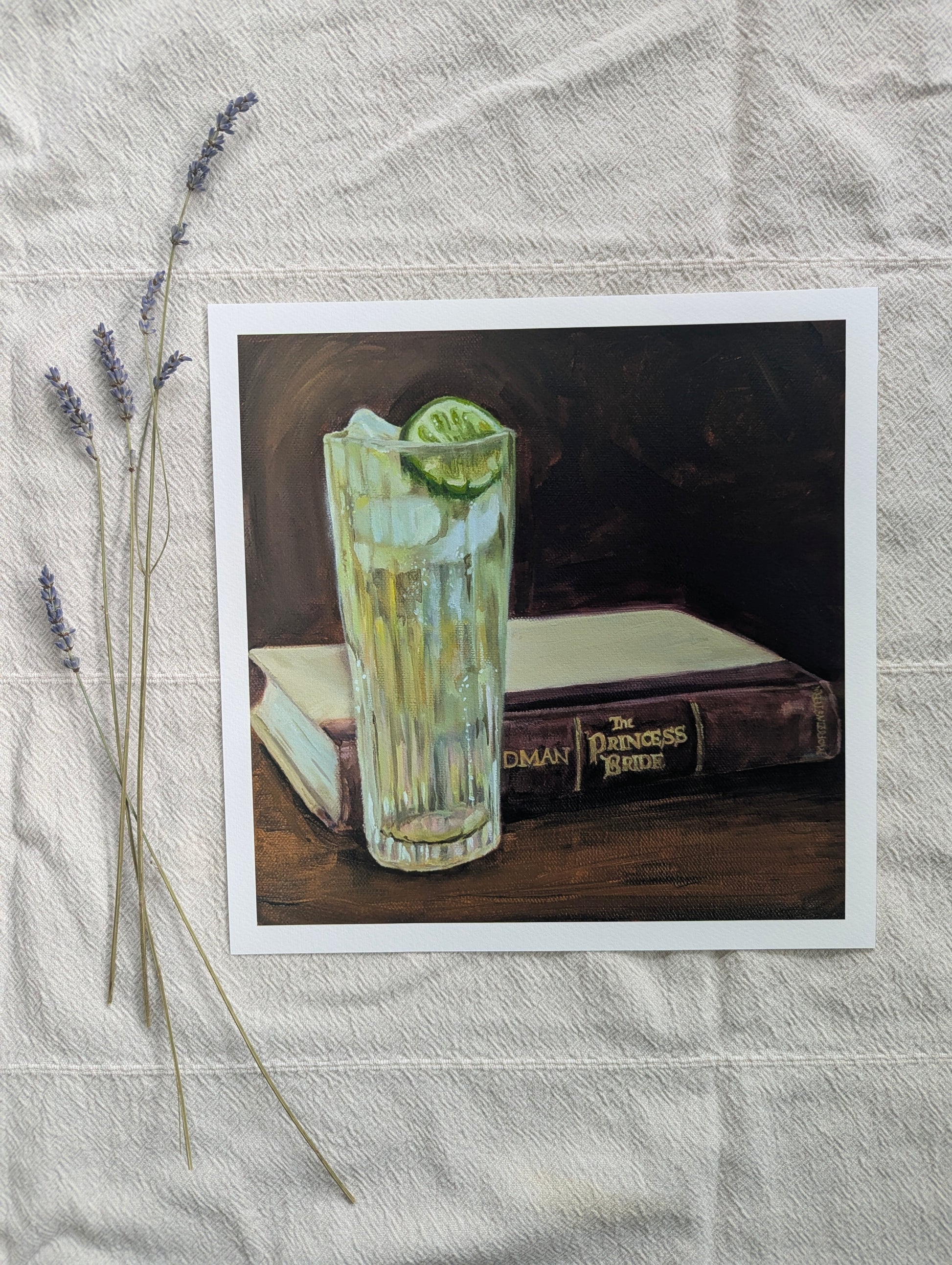 A square art print featuring a still life painting of a tall glass of gin and tonic with a lime wedge, set on a table next to a book, The Princess Bride,displayed on a textured white fabric with dried lavender sprigs.